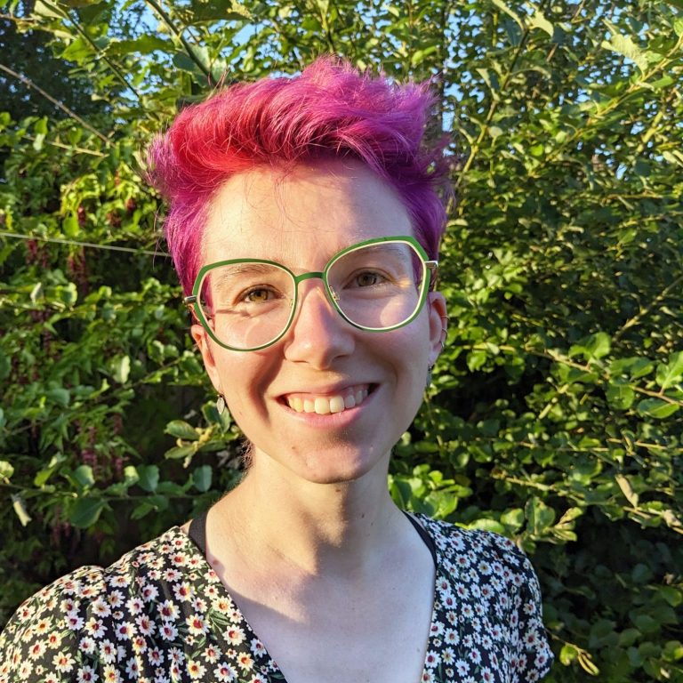 A photo of me smiling, I have pink hair and green glasses, there are trees in the background